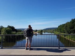 Thumbnail #Überlandflieger Ein Mann mit Rucksack steht auf einer Brücke und schaut auf einen Fluß