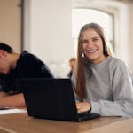 Mehrere junge Erwachsene sitzen in einem Klassenraum. Junge Frau im Vordergrund arbeitet am Laptop schaut lächelnd in die Kamera.